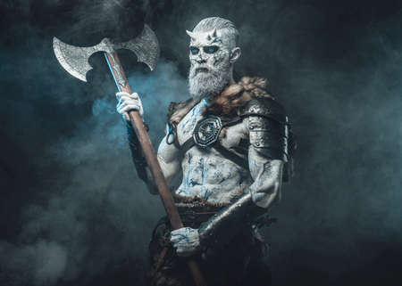 Risen from the dead northern warrior with horns and pale skin holding his two handed axe in frosty fog and dark background.の写真素材