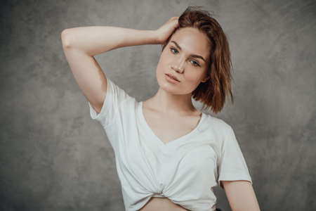 Brown haired beautiful woman dressed in white shirt with bare abdomen poses in gray background holding her head.の写真素材