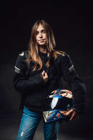 Female motorcyclist dressed in black jacket with jeans poses in dark background holding protective...
