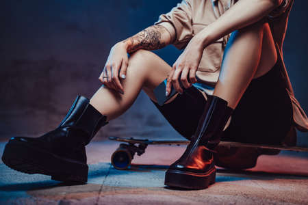 Portrait of female model with tattoo and dressed in stylish clothing she sits on skate holding smartphone in dark background.の写真素材
