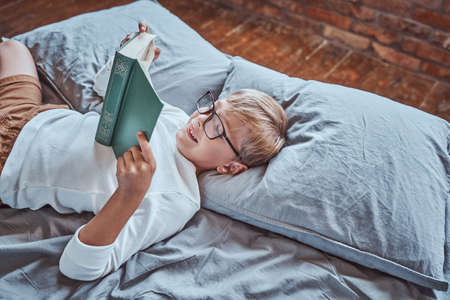 Dressed in casual clothing cheerful and smiley boy reads book lying down on a bed in living room.の写真素材