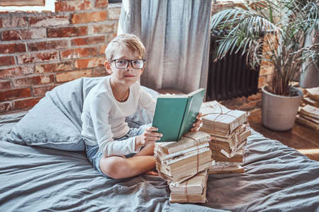 Clever and cheerful preschool child with glasses reads a book sitting on a bed there are many of books in living room.の写真素材