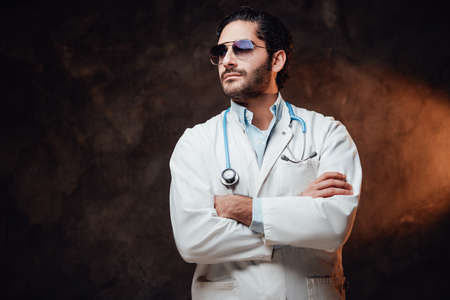 Self confident and brutal doctor with sunglasses in white labcoat poses with crossed arms in dark background.の写真素材