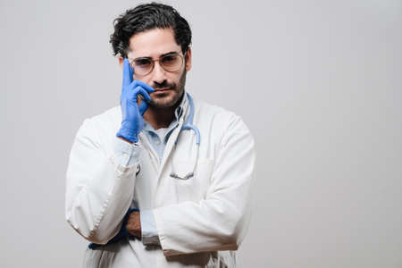Confident and skilled doctor dressed in white coat with rubber gloves and weared with glasses poses in white background.の写真素材