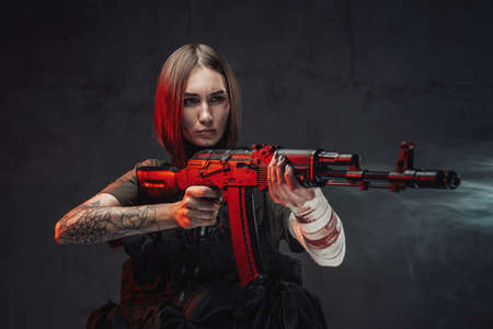 Dressed in black armour and dark shirt seductive martial woman with short haircut aims with rifle in...