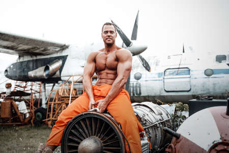 Positive athlete in orange clothing looking at camera and sitting on a engine of airplane in daytime.の写真素材