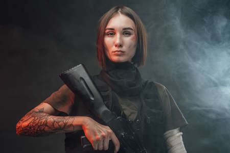 Seductive and tattoed woman dressed in modern and military body armour holding assault rifle in dark...