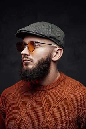 Headshot of bearded hipster posing in dark background weared with glasses and cap.