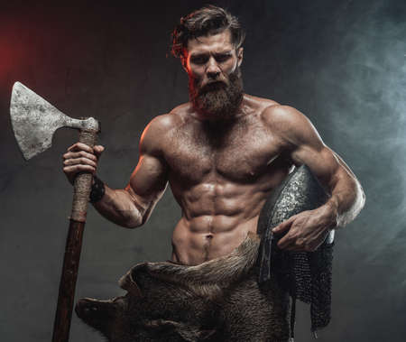 Serious and muscular viking with beard and naked torso staying in dark background with lights and smoke holding helmet and axe.の写真素材