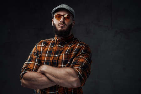 Dressed in checkered shirt with cap and weared with sunglasses bearded hipster guy poses in dark background with crossed arms.の写真素材