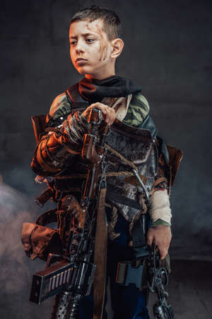 Armed with pistol and riffle apocalyptic child with blood on his face and dressed in rags stays in smokey background.の写真素材