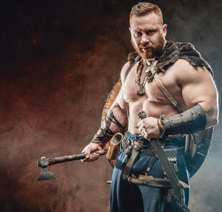 Shirtless and handsome scandinavian warrior with muscular build holding hatchet and knife in smokey background.の写真素材