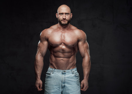 Caucasian topless guy with bald head and muscular build in jeans shorts poses in dark background looking at camera.の写真素材