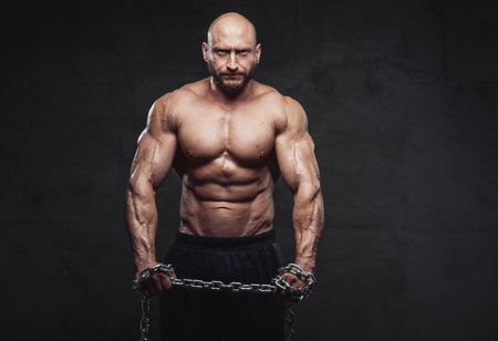 Strong and sportive man with muscular topless build poses holding heavy steel chains in dark background looking at camera with serious face.の写真素材