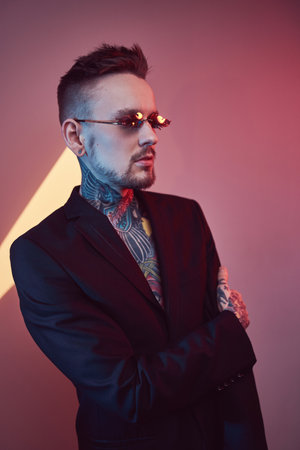 Portrait of a tattooed and fashionable hipster guy with stylish hairstyle in black suit weared with sunglasses poses in colourful background.の写真素材