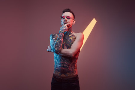 Shirtless and bearded bearded and tattooed guy with stylish hairstyle and sunglasses poses in colourful background with lights.の写真素材