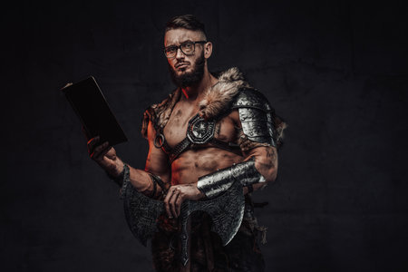 Holding a book scandinavian warrior weared with glasses dressed in light armour with two handed axe in dark background.の写真素材