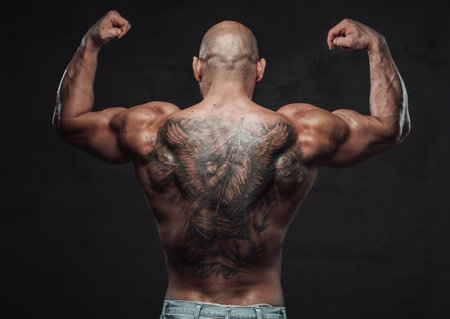 Shirtless and powerful bodybuilder poses in dark background with raised hands showing his muscular and tattooed back.の写真素材