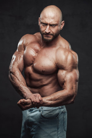 Bearded and hairless man with powerful and muscular build in jeans shorts poses with angry face in dark background looking at camera.の写真素材