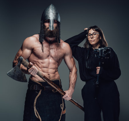 Muscularbearded guy with naked torso weared with helmet holding axe and photographer with glasses in...