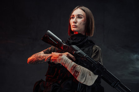 Dressed in dark armour military woman with short haircut and bandaged hand holds her assault rifle in dark smokey background.の写真素材
