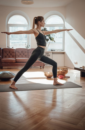 Dressed in black sportswear caucasian woman works out doing yoga staying on carpet with outstretched arms in shiny living room.の写真素材