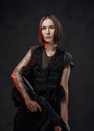 Beautiful woman with short haircut dressed in black armour with dark shirt holding assault rifle in dark background.の写真素材