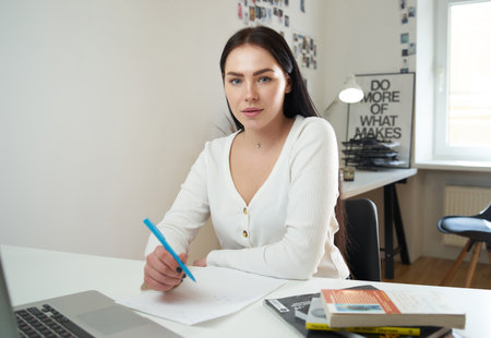 Caucasian female journalist with black hairs dressed in white shirt sits at table looking at camera...