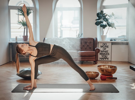 Graceful and beautiful sportswoman in black clothing works out doing yoga on carpet in atmospheric living room with sunbeams.の写真素材