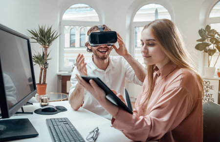 Bearded man in white shirt uses virtual headset and smiles while his female assistant with long brown hairs works on tablet in office room.の写真素材