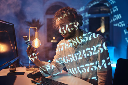 Working late office worker with beard and curly hairs looks at his mobile phone sitting at table with desk computer in dark room.の写真素材