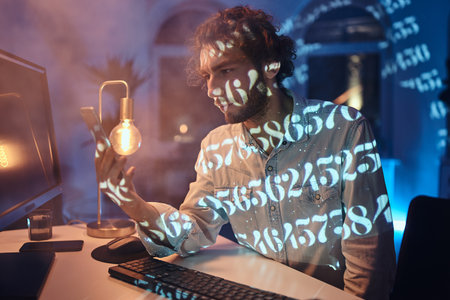Bearded caucasian worker with curly hairs sits at table looking at his telephone in dark room with...