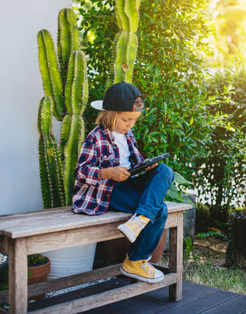 Relaxed kid dressed in casual clothing sits on bench and plays some games on his tablet in park.の写真素材