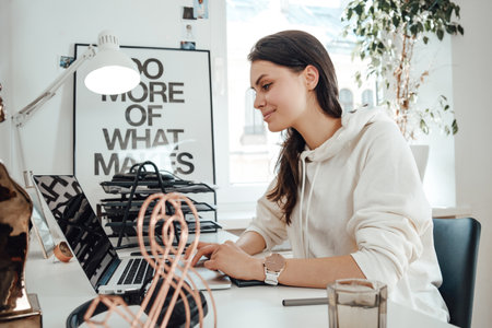 Young female business person works on laptop and smiles sitting at table in comfortable office.
