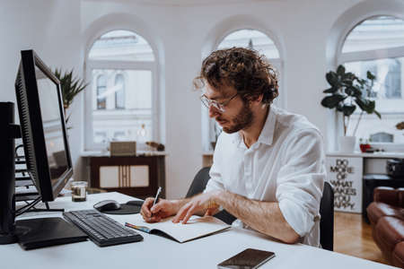 Serious caucasian guy writes on notebook sitting at table there is his computer in modern office.の写真素材