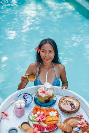 Asian woman and asian food. Young woman with dark hairs poses in swimming pool with floating table and cup of juice.の写真素材