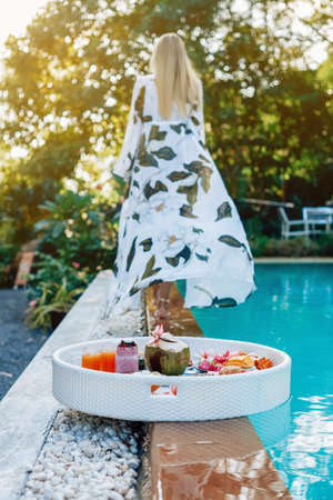 Floating table full filled with fresh and different tropical food and drinks in background of swimming pool and woman going away.の写真素材