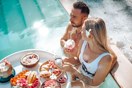 Portrait of succesful sweet couple on their vacations in Thailand. Woman and man drink cocktails and eat asian food in swimming pool together.の写真素材