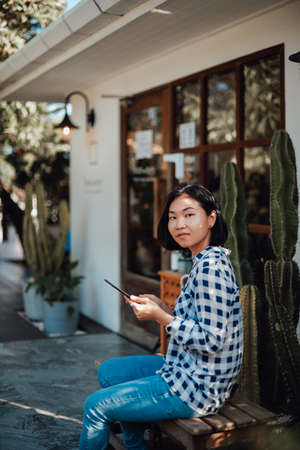 Portrait of a young asian woman she dressed in jeans with shirt and sitting on a bench using her telephone outside.の写真素材