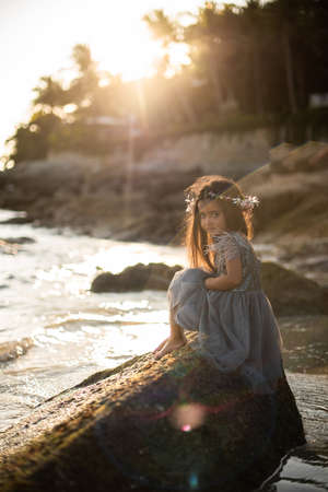 Portrait of journey of a little girl in thailand. Preteen girl in dress poses sitting on the rock barefot looking at the camera in the sunshine.の写真素材