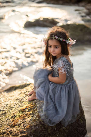 Portrait of journey of a little girl in thailand. Preteen girl in dress poses sitting on the rock barefot looking away in the sunshine.の写真素材
