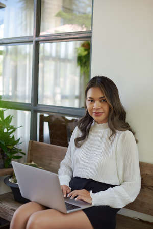 Cute and joyful asian girl dressed in casual clothing sits on bench outside working on laptop and looking at camera.の写真素材