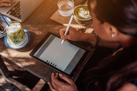 Warm portrait of a asian woman she sits at table with coffee and water cups and works on her tablet outside in sunny day.の写真素材