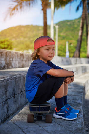 Cheerful child dressed in sportswear with baseball cap sits on skateboard looking away. Vacations and relaxation in Thailand.の写真素材
