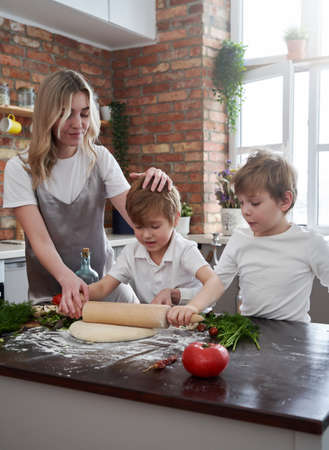 Cheerful and joyful boys and their young mother cook a pizza and learn to cook in kitchen in...