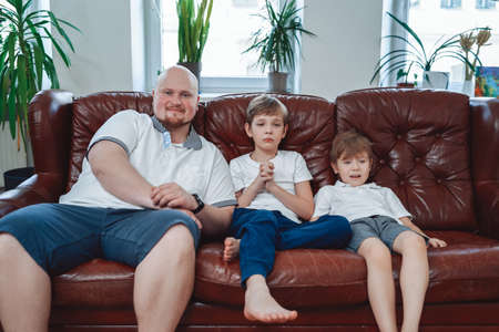 Fatherhood and two little boys. Mature and bald man with his children sits on sofa and pose on camera in modern apartment.の写真素材