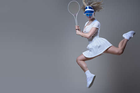 Caucasian female tennis player dressed in white sportswear with blue cap poses in white background jumping and holding racquet.の写真素材