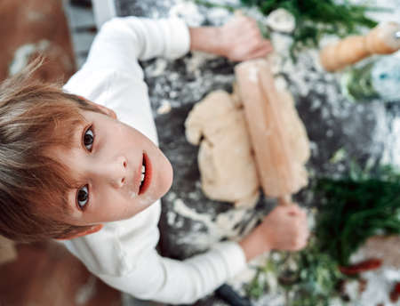 Portrait of a young and astonished boy in white shirts he looks at camera and rolls a dough...