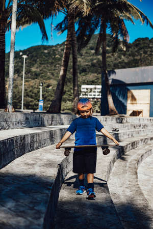 Daytime pastime and sports hobby. Youth boy in cool clothing with cap walking through stairs in park holding a skateboard outdoors.の写真素材