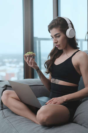 Caucasian woman in black sportswear with headphones sits on sofa using laptop and holding junk food...